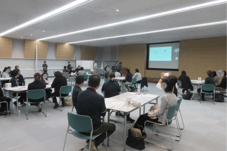 広島県三次市で開催された8050問題に関する合同研修会。各グループの代表者が検討内容を発表し、会場の参加者が耳を傾けている様子。講師はZoomを通じて発表を共有し、講評を行った。